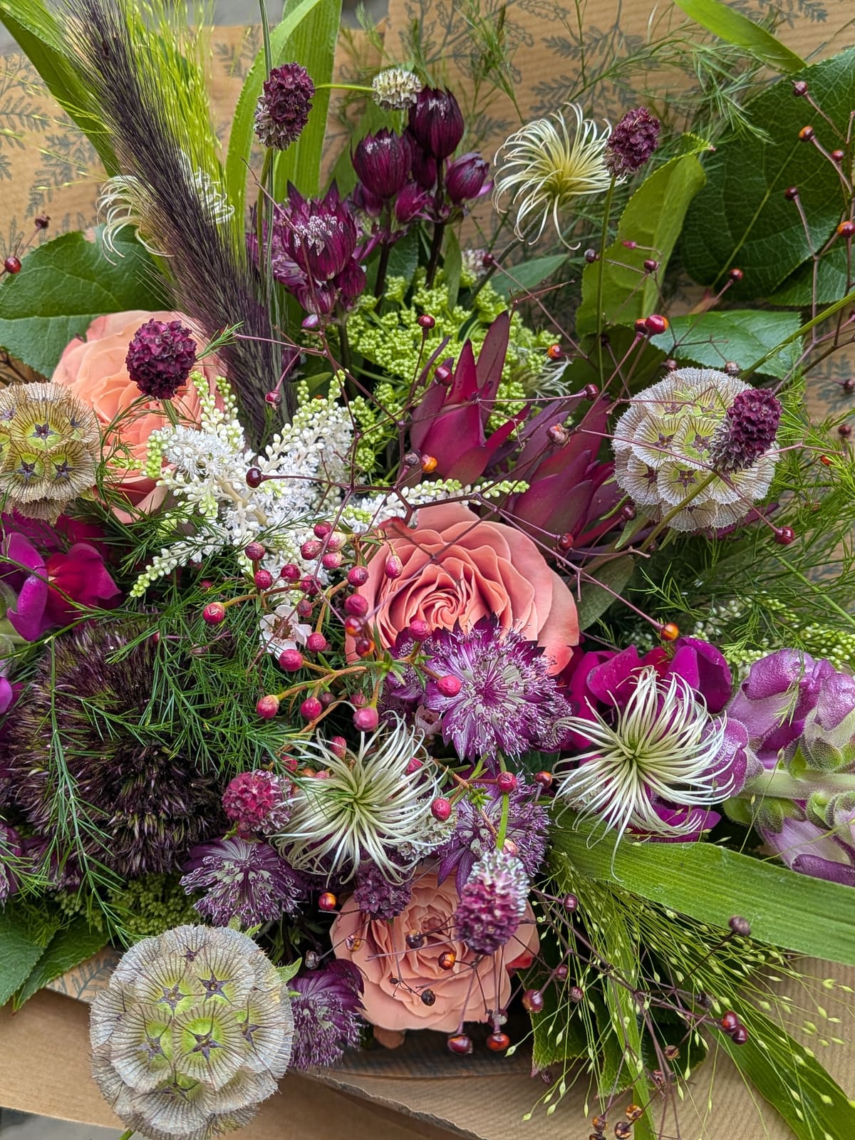 A wild hand-tied bouquet of coral roses, plum astrantia, scabiosa seedheads and grasses in patterned kraft paper