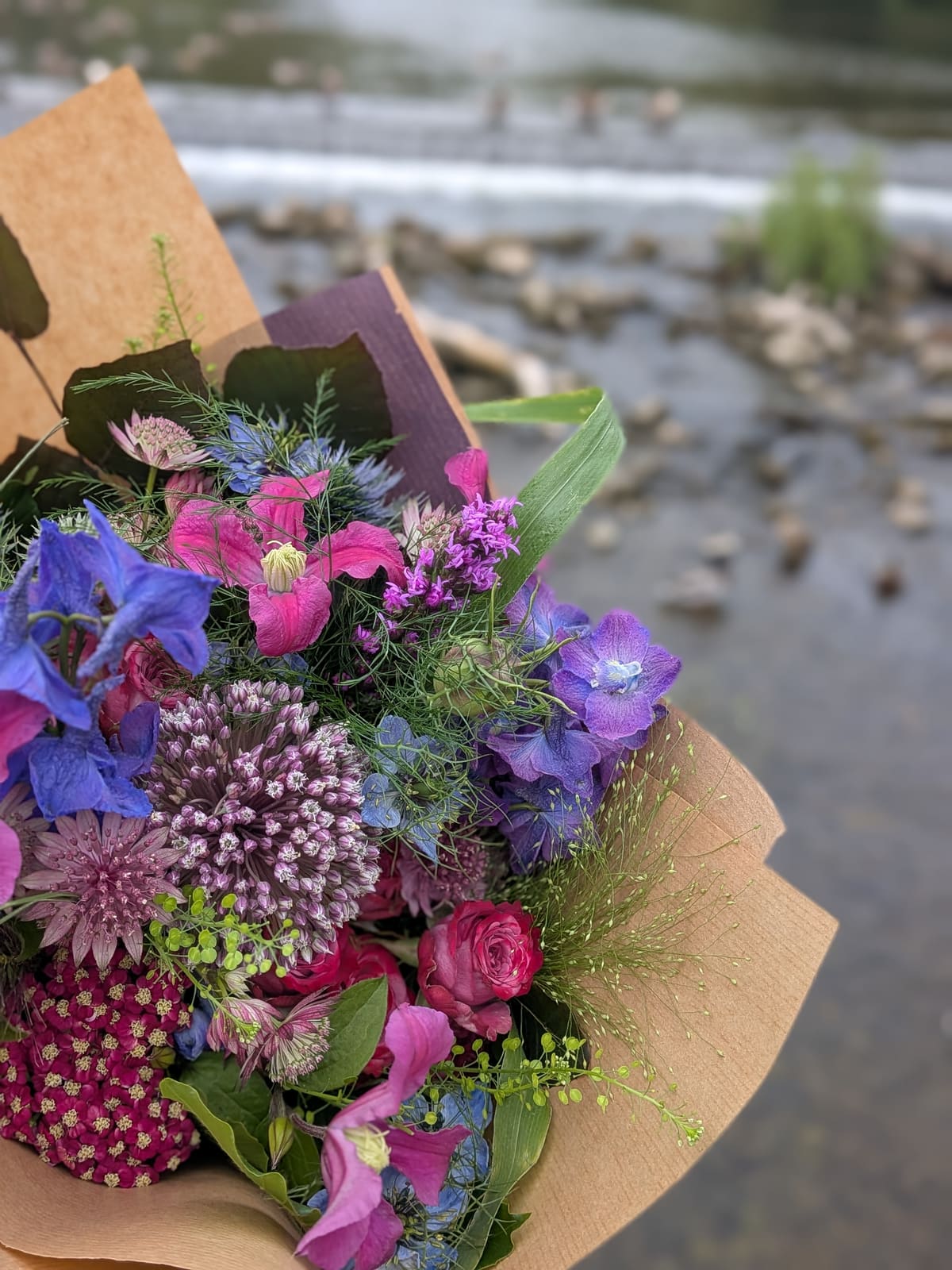 A jewel-toned bouquet of delphinium, clematis, allium and roses held above the river weir in Bakewell