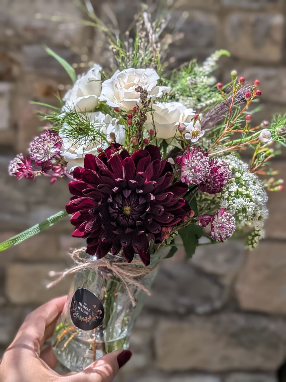 A hand-tied posy of burgundy dahlia, white roses and astrantia in a glass jar with The Bramble Hollow label, held against a stone wall